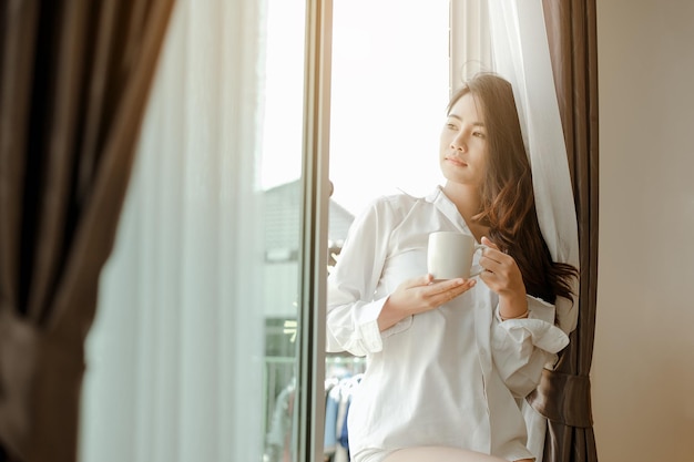 Asia mujer joven que vive en casa relajarse y beber una taza de café caliente en el dormitorio de vacaciones. Asia, asia, relajarse, solo, tecnología, concepto de estilo de vida.