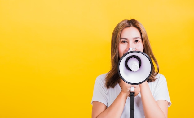 Asia tailandesa feliz retrato hermosa linda mujer joven de pie para hacer un mensaje de anuncio gritando en megáfono mirando a la cámara, tiro de estudio aislado sobre fondo amarillo con espacio de copia