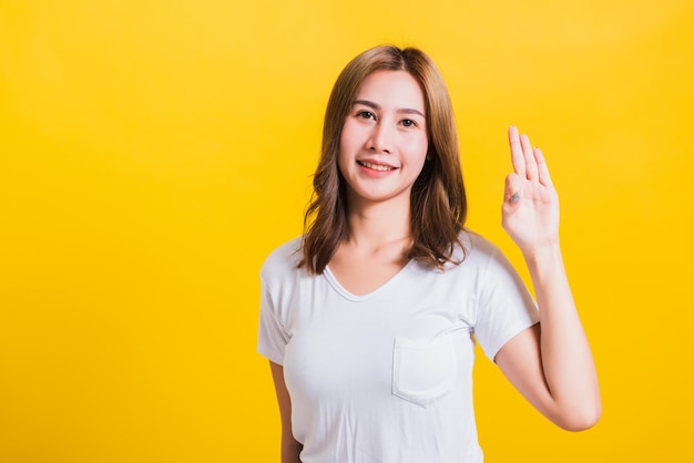 Asia tailandesa feliz retrato hermosa linda mujer joven de pie usando una camiseta que muestra el signo de ok con los dedos mirando a la cámara, estudio aislado filmado sobre fondo amarillo con espacio de copia