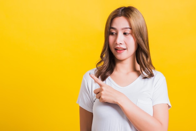 Asia tailandesa feliz retrato hermosa linda mujer joven de pie usar camiseta blanca apuntando con el dedo hacia el lado mirando hacia el otro lado, tiro del estudio aislado en fondo amarillo con espacio de copia