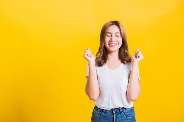 Asia tailandesa feliz retrato hermosa linda mujer joven de pie usar camiseta hace puños levantados celebrando su éxito ganador ella cierra los ojos aislado, estudio filmado sobre fondo amarillo con copia spa