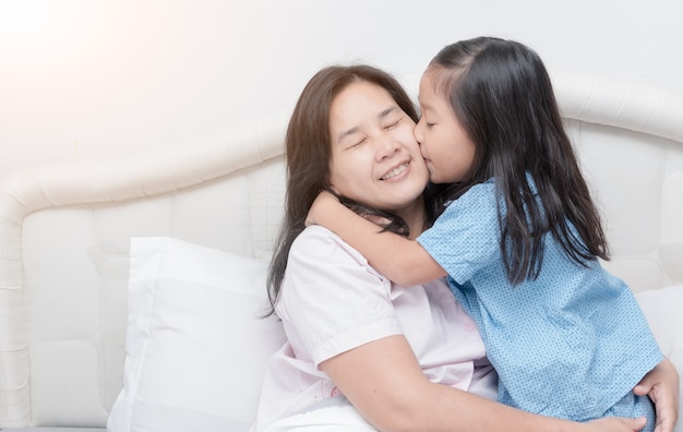 Asiática doce menina está abraçando e beijando sua bela jovem mãe na bochecha enquanto está sentado na cama