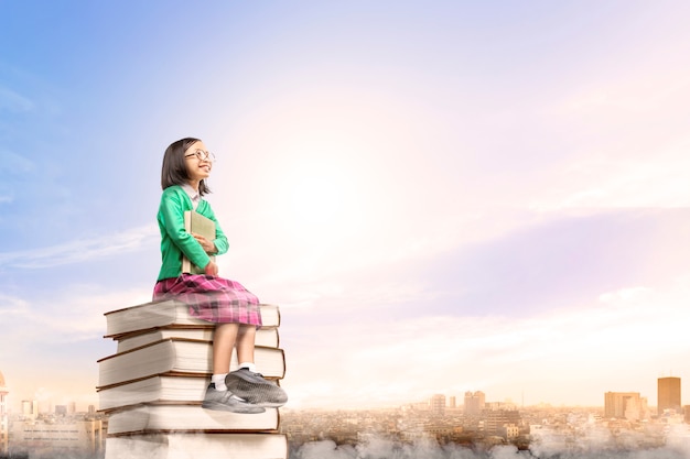 Foto asiática linda garota de óculos, segurando o livro enquanto está sentado na pilha de livros com a cidade e o céu azul