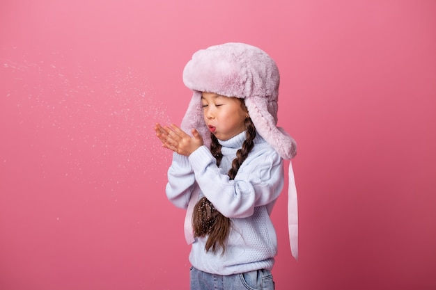 Asiática menina bonitinha com roupas de inverno sopra neve das palmas das mãos. Fim do inverno, fundo rosa, espaço para texto