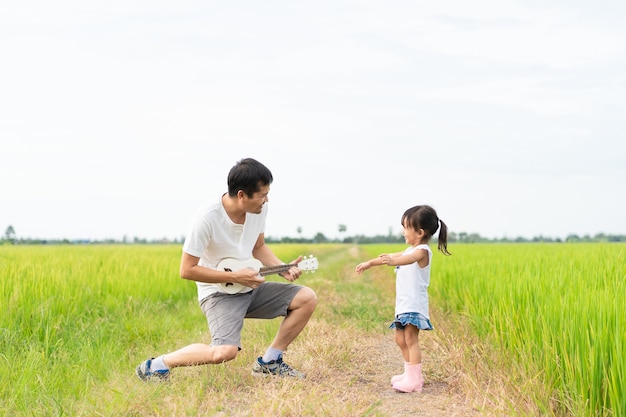 Foto asiática pai e filha estão tocando o ukulele e dançando juntos no campo de arroz