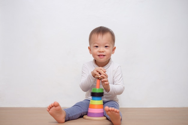 Asiática pequena bonito 18 meses / 1 ano de idade bebê criança brincadeira com brinquedo pirâmide de madeira colorida / brinquedo anel de empilhamento. Criança brincando com brinquedo educacional isolado na parede branca, com espaço de cópia