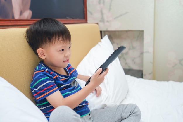 Asiático de 3 a 4 años de edad, niño pequeño, niño sentado en la cama con el control remoto del televisor y viendo la televisión en la habitación de su casa