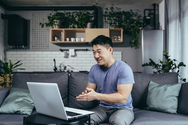 Asiático en casa hablando en video usa una computadora portátil mira la pantalla hombre en casa sentado en el sofá