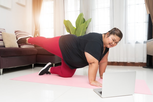 Asiático de mulher gorda se exercitando e está usando um computador laptop em casa, o conceito de ideia de esporte e recreação.