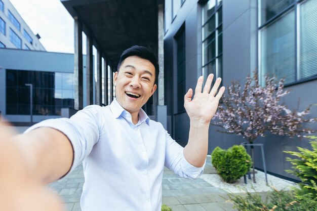 Asiático falando em videochamada usa telefone olha para a câmera e acena alegremente com as mãos gesto de saudação