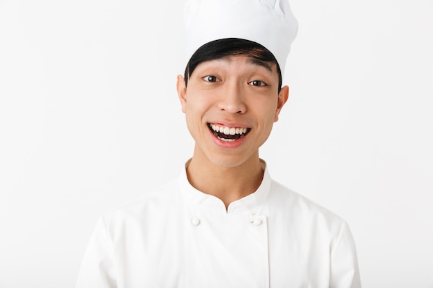 Asiático feliz chefe homem com uniforme de cozinheiro branco, sorrindo para a câmera em pé, isolado sobre uma parede branca