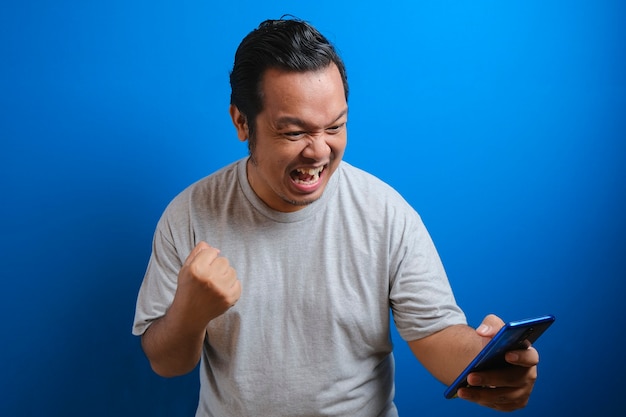 Un asiático gordo con una camiseta gris se ve feliz por las buenas noticias que recibió de su teléfono inteligente. Los hombres muestran gestos de victoria. fondo azul