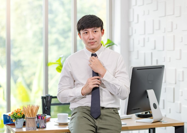 Asiático joven apuesto profesional exitoso hombre de negocios empleado en camisa de negocios formal ajustando corbata sentado apoyado en el escritorio de trabajo lleno de papelería de monitor de computadora y taza de café.