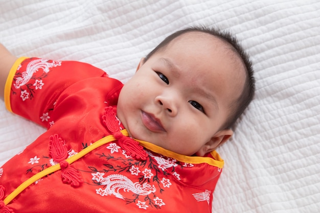 Asiático lindo bebé niño traje chino Cheongsam niño pequeño acostarse en la cama en casa sonriendo riendo de buen humor, curiosidad infantil chino niño niño mirando algo, feliz año nuevo chino concepto