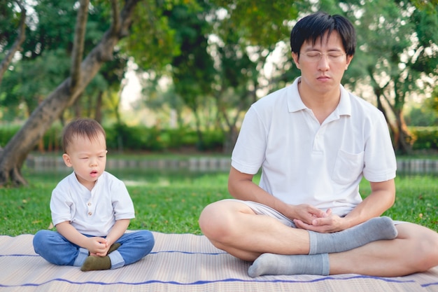 Asiático pai com os olhos fechados e criança de menino de 1 ano de idade criança pratica ioga &amp; meditando ao ar livre na natureza no verão, conceito de estilo de vida saudável