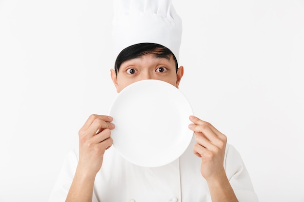 asiático satisfeito chefe com uniforme branco de cozinheiro, sorrindo para a câmera enquanto segura um prato isolado na parede branca