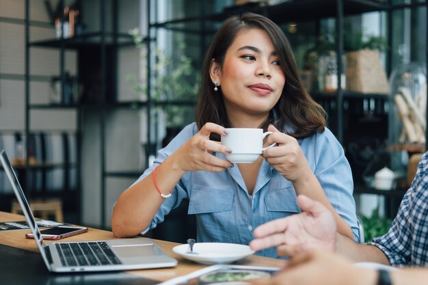 Asiatin im blauen Hemd in trinkendem Kaffee des Cafés und in der Unterhaltung mit Freundlächeln und glücklichem Gesicht