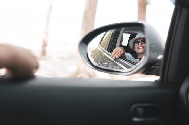 Asiatin im weißen Hemd untersucht den Seitenansichtspiegel und lächelt beim Sitzen in ihrem Auto, Reisekonzept.