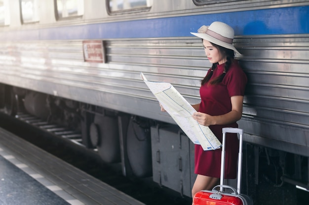 Asiatin schwanger im roten Kleid, das rotes Gepäck und Blick auf die Karte auf Eisenbahn trägt