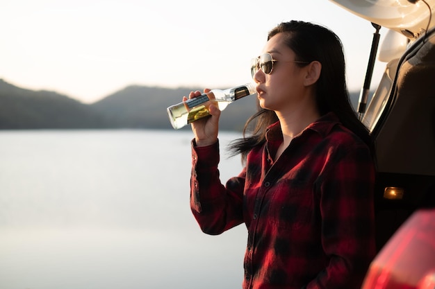 Asiatin trinkt Bier auf der Rückseite eines Autos im Hintergrund eines Flusses und eines Berges