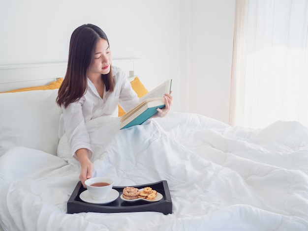 Asiatinlesebuch und trinkender Kaffee auf Bett morgens