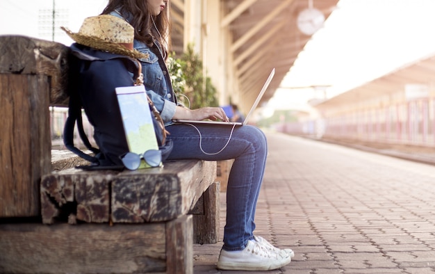 Asiatinreisender mit Rucksack, Hut, Karte, Sonnenbrille, Kopfhörer und Anwendung des Laptops an der Bahnstation in den Feriensommerferien