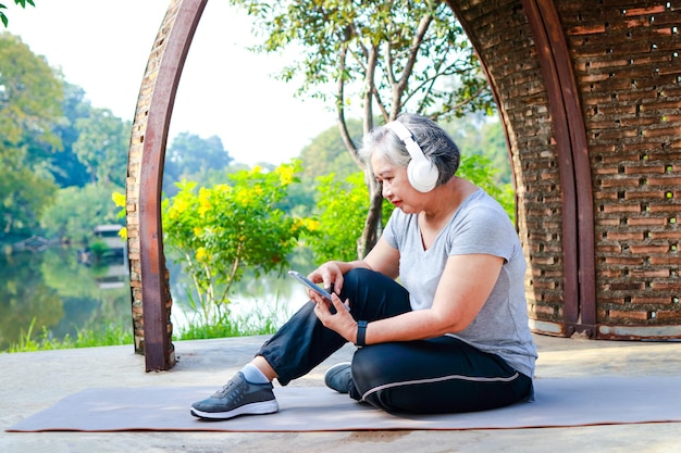 Asiatische ältere Frau, die morgens im Park trainiert Sie sitzt und entspannt sich, hält ihr Smartphone und trägt Kopfhörer, um Musik zu hören. konzept der alten gesundheitsversorgung