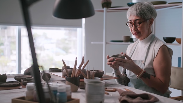 Asiatische ältere Frau, die zu Hause Töpferarbeiten genießt Eine Keramikerin stellt in einem Studio neue Töpferwaren her