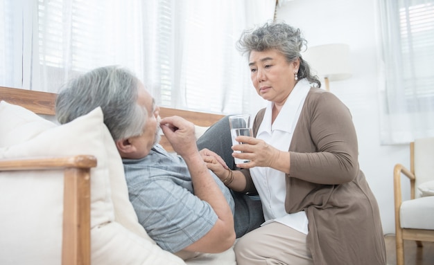 Asiatische ältere Frau kümmern sich darum, dem älteren Mann, der Medikamente nimmt und Wasser trinkt, ein Glas Wasser zu geben, während er zu Hause auf der Couch liegt