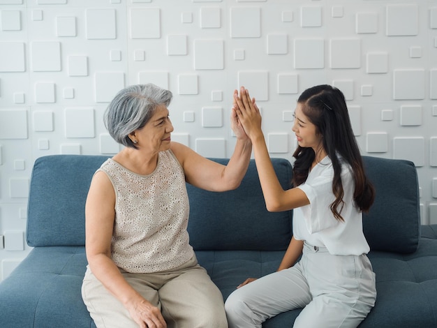 Asiatische ältere Frau Mutter gibt ihrer fröhlichen Tochter High Five junge Schönheit weiblich sitzt auf grauem Couchsitz im weißen Raum Mutter und Tochter feiern den Sieg gewinnen zusammen und klatschen in die Hände