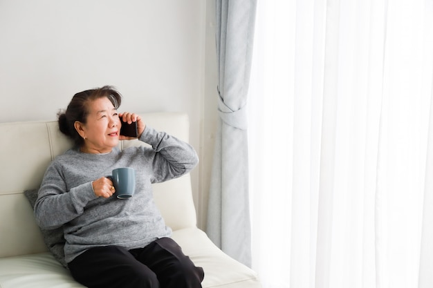 Asiatische ältere Frauenmutter, die mit Handy spricht und Kaffee im Wohnzimmer trinkt