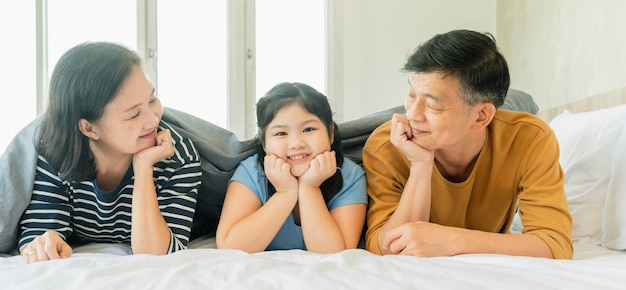 Asiatische ältere Großeltern, die mit Enkelkindern ein hübsches weibliches Kind auf dem Bett im Schlafzimmer legen, glückliche Familie, die zusammen im Bett spielt