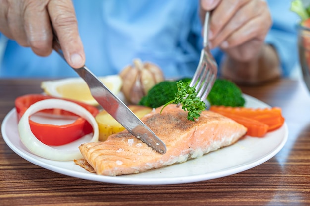 Asiatische ältere oder ältere alte Dame Patientin, die Lachssteakfrühstück mit gesundem Gemüseessen isst, während sie im Krankenhaus sitzt und hungrig auf dem Bett sitzt.