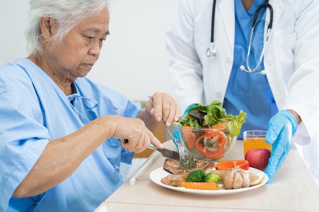 Asiatische ältere oder ältere Patientin der alten Dame, die hoffnungsvoll und glücklich Frühstück und gesunde Gemüsekost isst, während sie im Krankenhaus sitzt und hungrig auf dem Bett sitzt