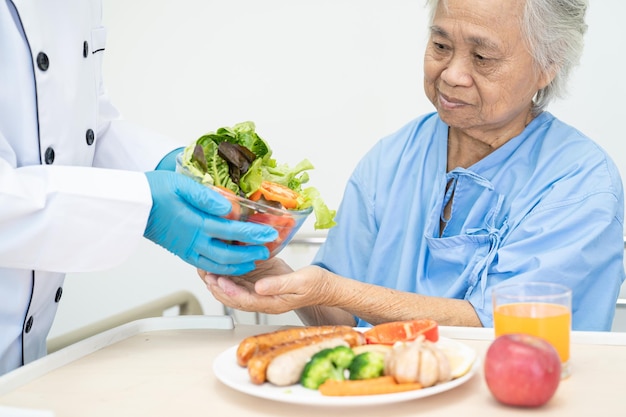 Asiatische ältere oder ältere Patientin der alten Dame, die hoffnungsvoll und glücklich Frühstück und gesunde Gemüsekost isst, während sie im Krankenhaus sitzt und hungrig auf dem Bett sitzt