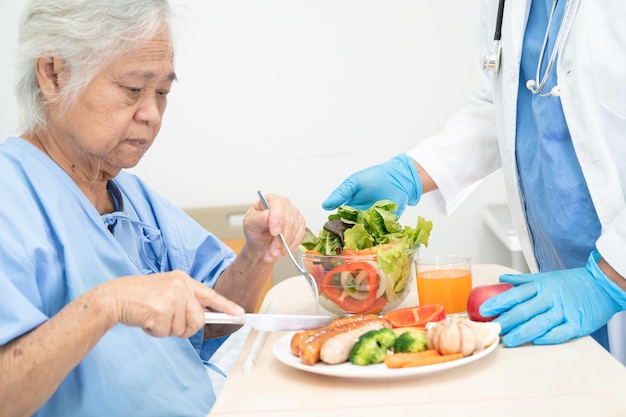 Asiatische ältere oder ältere Patientin der alten Dame, die hoffnungsvoll und glücklich Frühstück und gesunde Gemüsekost isst, während sie im Krankenhaus sitzt und hungrig auf dem Bett sitzt