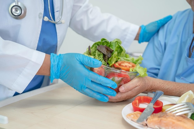 Asiatische ältere oder ältere Patientin der alten Dame, die hoffnungsvoll und glücklich Frühstücksgemüse isst, während sie im Krankenhaus sitzt und hungrig auf dem Bett sitzt