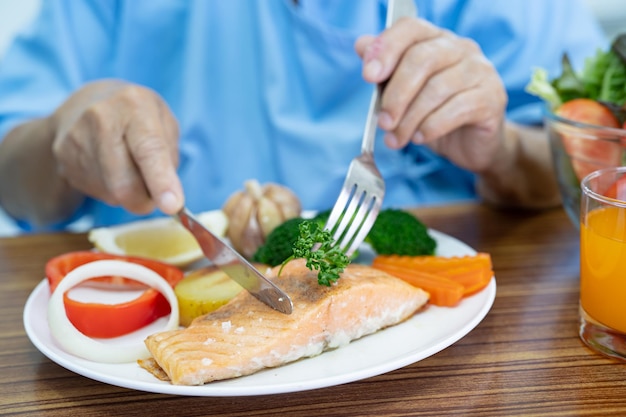 Asiatische ältere oder ältere Patientin der alten Dame, die Lachssteak-Frühstück mit pflanzlicher gesunder Nahrung isst, während sie im Krankenhaus sitzt und hungrig auf dem Bett sitzt