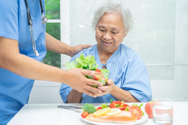 Asiatische ältere Patientin isst im Krankenhaus Lachsbechsteak zum Frühstück mit Gemüse und gesundem Essen