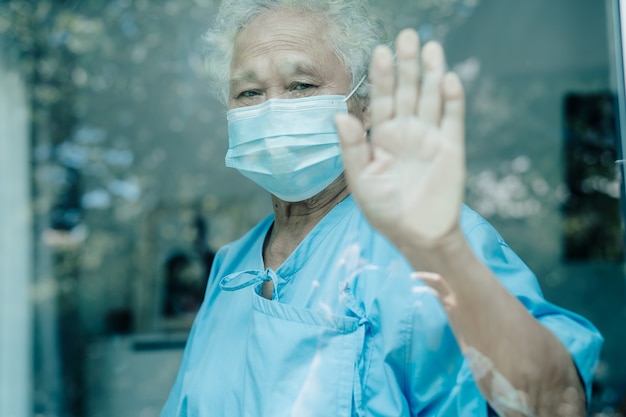 Asiatische ältere Patientin mit Maske, die während des Coronavirus aus dem Fenster schaut, um in Quarantäne zu gehen