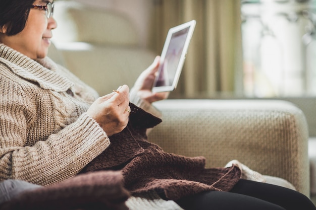 Asiatische alte Frau, die Häkeln mit Tablette zu Hause strickt