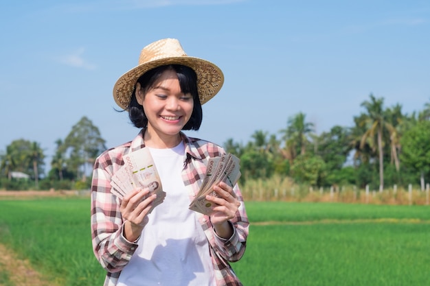 Asiatische Bauernfrau, die thailändisches Banknotengeld mit Lächelngesicht an grüner Reisfarm hält