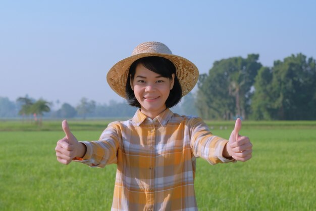 Asiatische Bauernfrauen tragen einen gelben Hemddaumen nach oben und lächeln auf der grünen Farm.