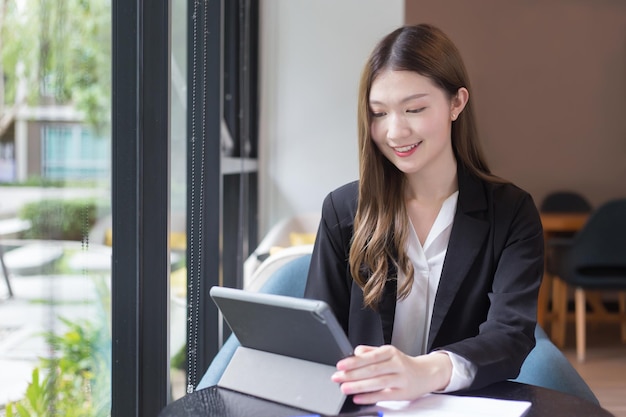 Asiatische berufstätige frau im schwarzen anzug arbeitet an einem tablet auf dem tisch im büro und arbeitet zu hause