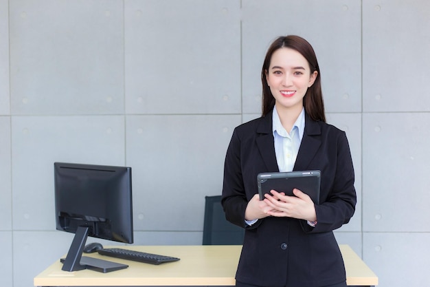 Asiatische berufstätige Frau im schwarzen Anzug hält Tablet in ihren Händen und lächelt selbstbewusst im Büro