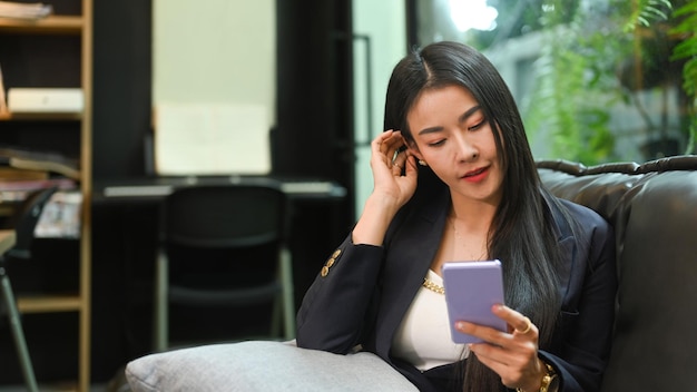 Asiatische berufstätige Frau mit Handy, während sie sich in ihrem persönlichen Büro auf dem Sofa ausruht