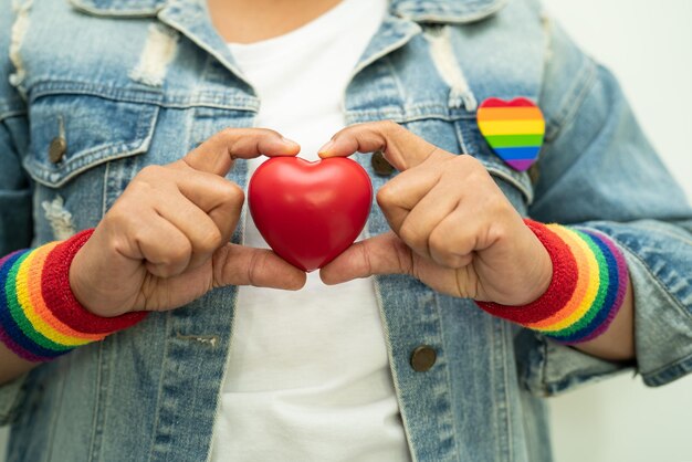 Asiatische Dame, die Armbänder mit Regenbogenfahne trägt und das rote Herzsymbol des LGBT-Stolzmonats hält, feiert jährlich im Juni soziale schwul-lesbische bisexuelle Transgender-Menschenrechte