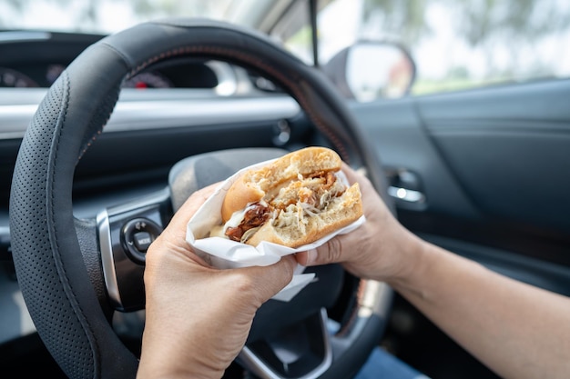 Asiatische Dame, die einen Hamburger zum Essen im Auto hält, ist gefährlich und riskiert einen Unfall