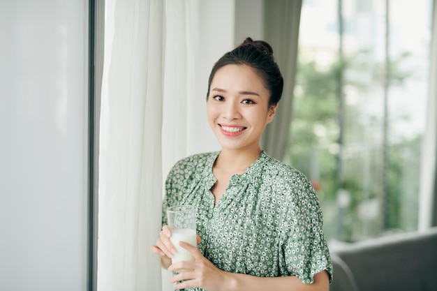 Asiatische Dame, die Glas Milch trinkt, während sie mit Kopierraum aus dem Fenster schaut