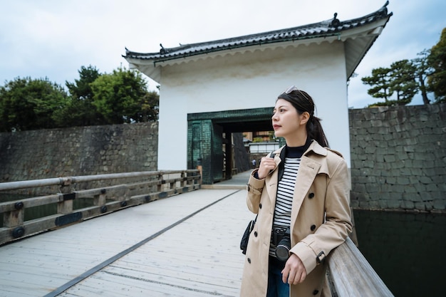 asiatische dame, die in die ferne schaut und sich auf die felsenarmlehne lehnt, macht eine pause auf einer brücke. Reisende, die ihren Arm auf dem Handlauf der Brücke ruhen lassen, genießen die Aussicht von der Grabenbrücke auf dem Gelände der Burg Nijo.